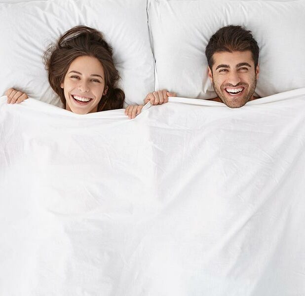 Glückliches Pärchen auf Ihrem Bett, beide ziehen sich die Bettdecke über den Kopf.